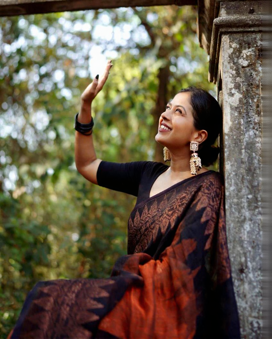 Rust orange Banarasi Silk saree with a black woven border featuring traditional motifs, worn by a smiling woman in an outdoor setting with concrete steps and greenery.