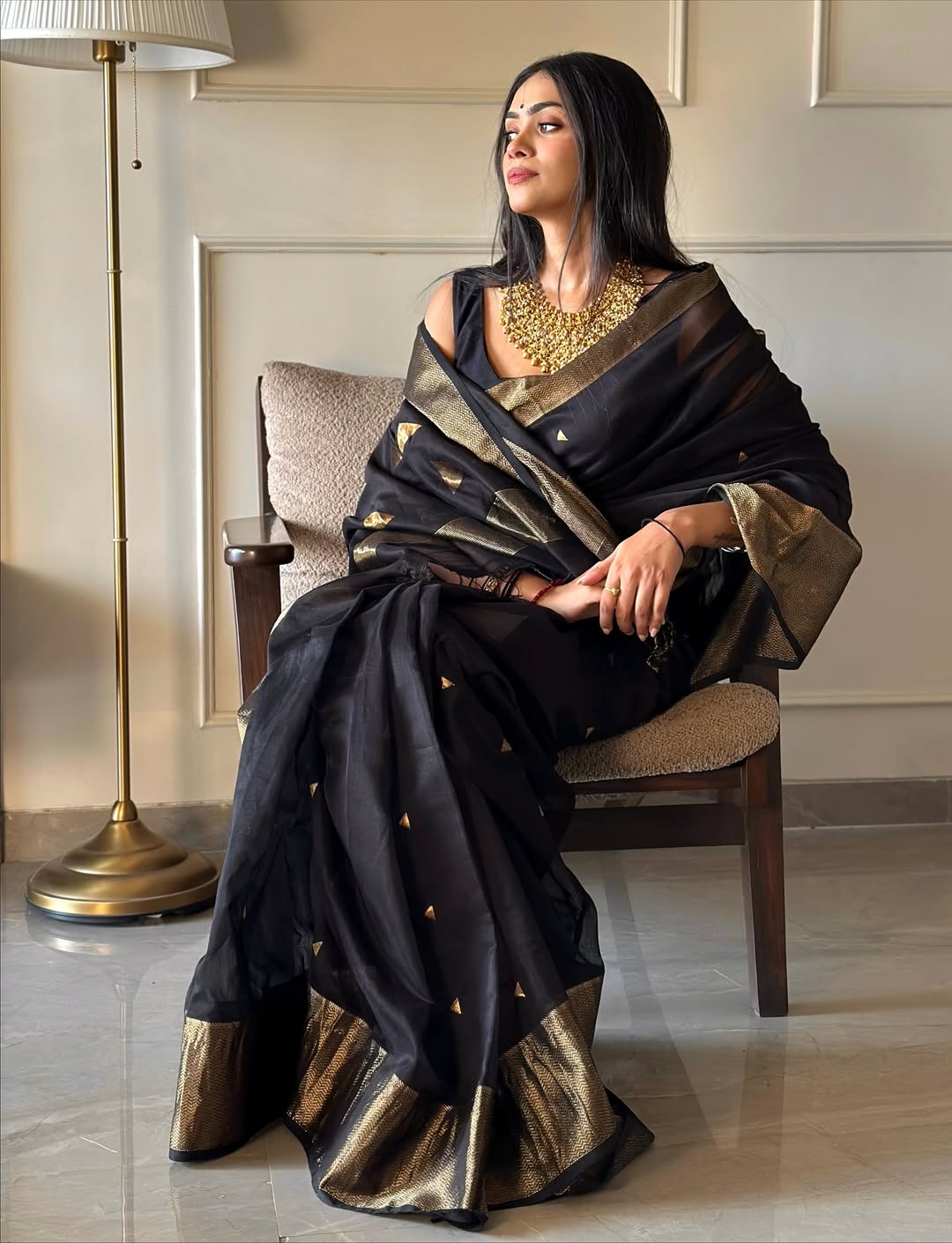 Woman in a black and gold saree sitting on a chair in a room.