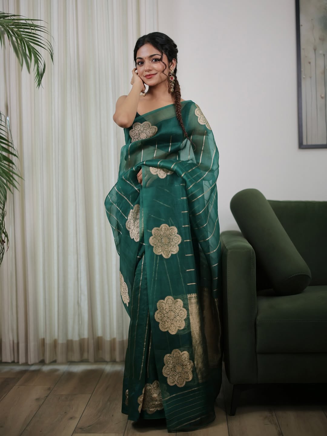 Woman in a green saree with gold patterns standing in a room with a couch and plant.