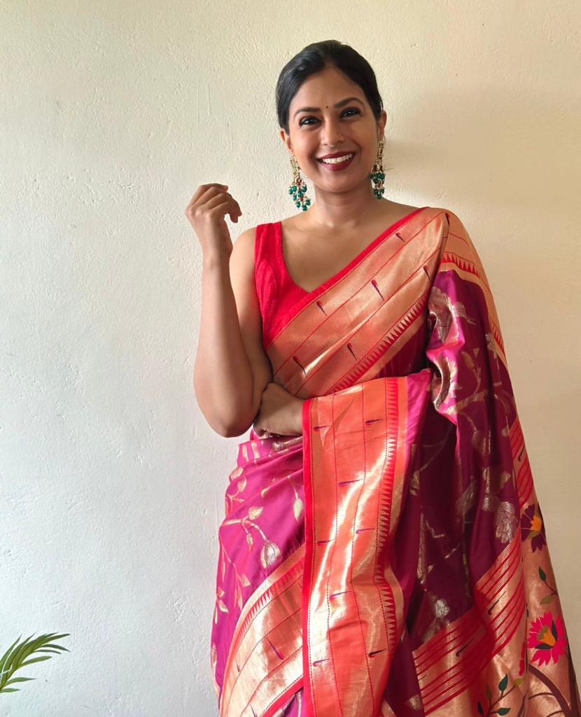 A smiling woman looking elegant in a vibrant rani pink Banarasi silk saree featuring a wide, lustrous gold brocade border and gold motifs. She pairs it with a simple red sleeveless blouse and statement green and gold earrings