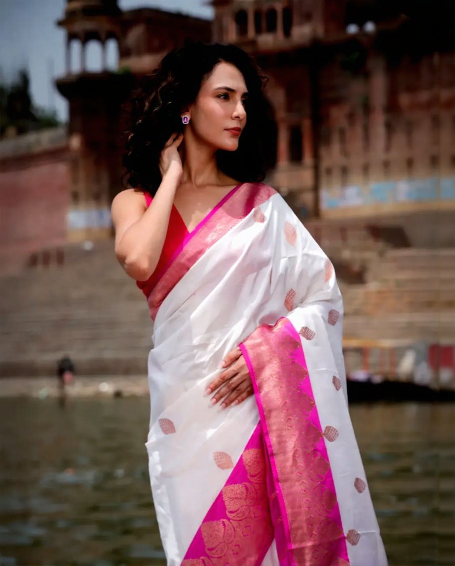 Woman in a white and pink saree standing by a body of water with historical architecture in the background