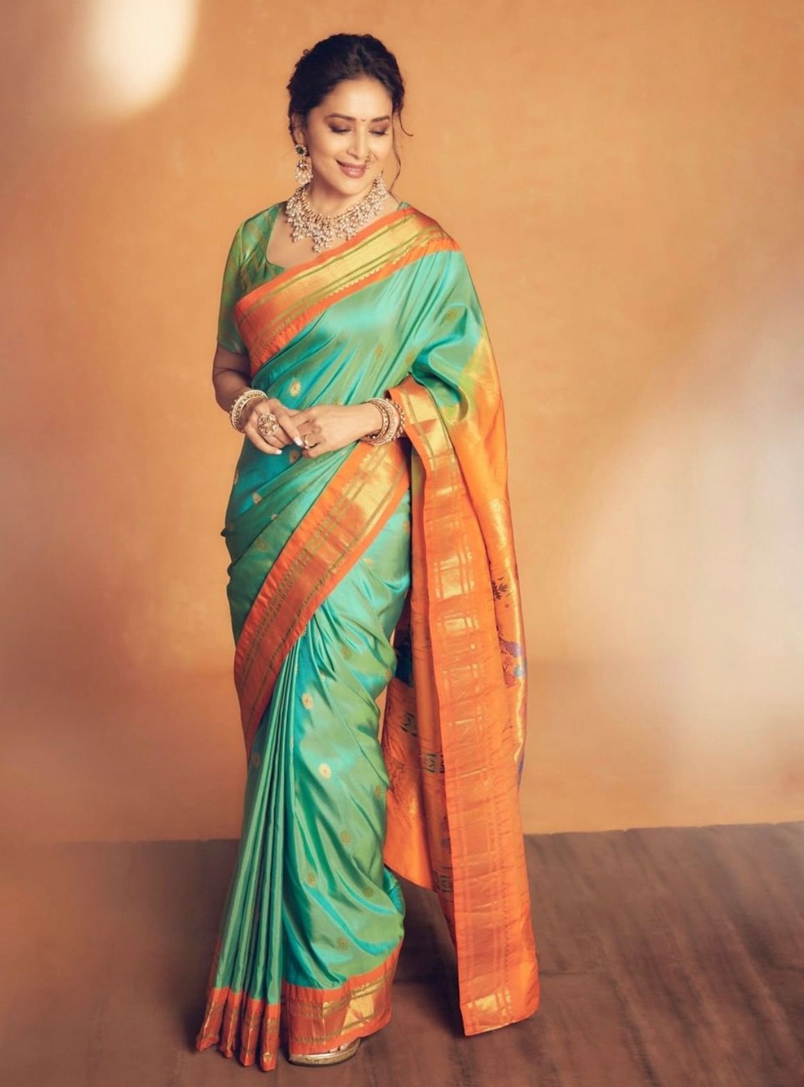 Luminous Soft Lichi Silk saree in teal/mint green with a wide, fiery orange contrast border featuring intricate temple zari weaving, gracefully worn by a smiling woman with traditional jewelry against an orange wall.