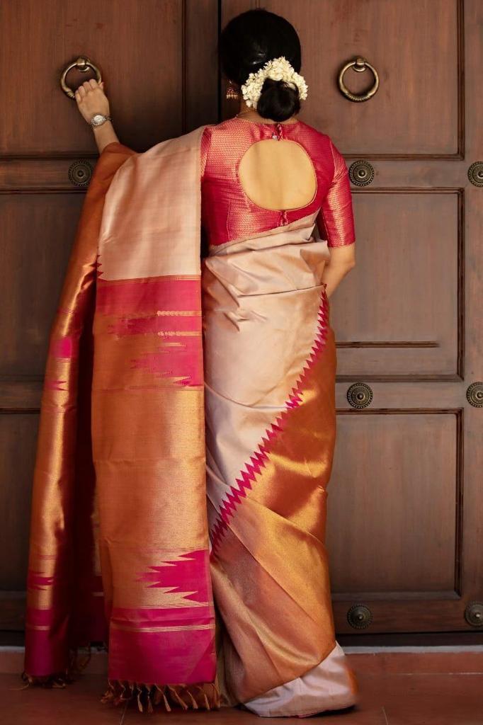 A smiling woman stands against a brick wall, elegantly draped in a soft Lichi silk saree in a blush golden-beige hue, featuring a rich rani pink and golden border, paired with a vibrant red blouse, gold temple jewelry, and gajra in her hair.