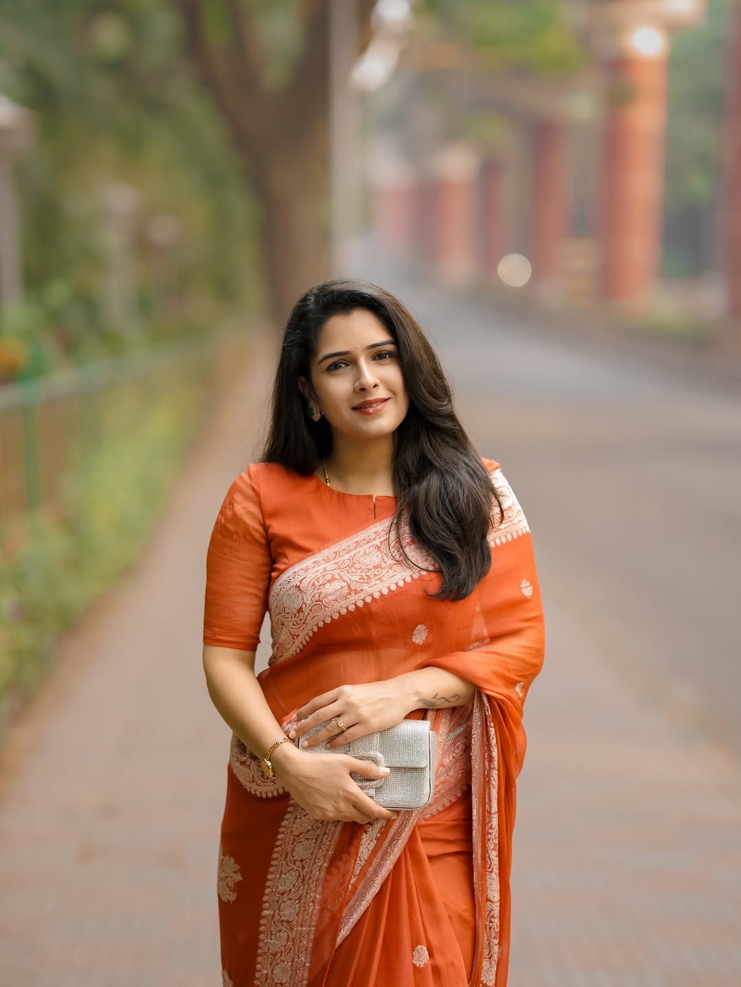 Woman in an orange saree standing on a path with greenery and a building in the background