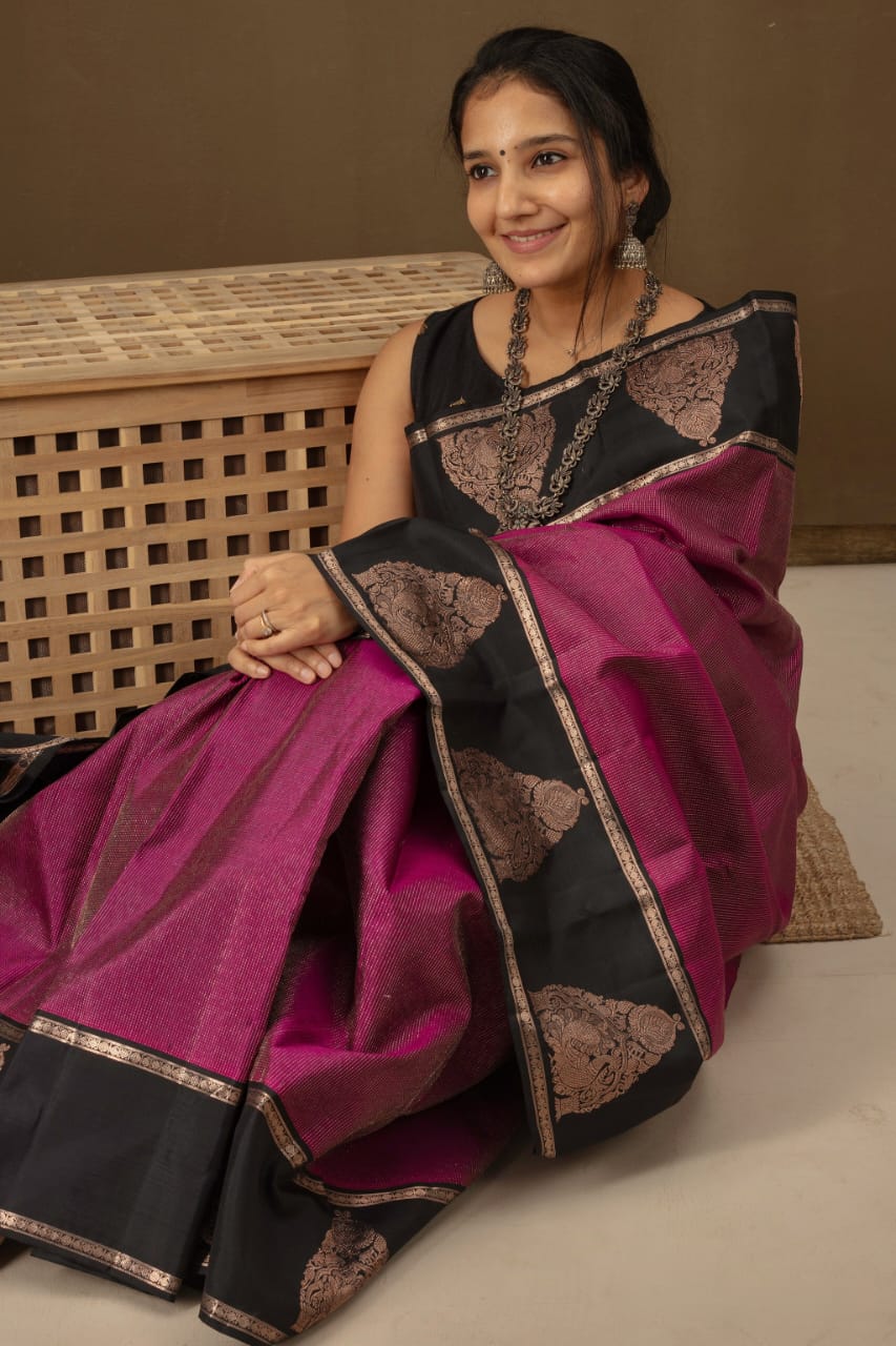 Deep fuschia/magenta Banarasi Silk saree with black contrast panels on the pallu and border, adorned with bronze zari paisley motifs, draped on a woman sitting on a wooden stool indoors.