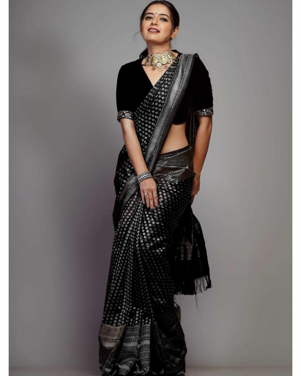 A woman models a black soft Lichi silk saree with silver patterned weaving, a matching velvet blouse, and traditional Indian jewelry, against a plain background.
