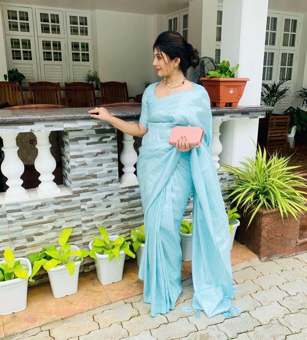 A woman stands gracefully on a porch, holding a pink clutch and wearing a soft, shimmering aqua blue Lichi silk saree with a matching blouse, accessorized with a delicate gold necklace.