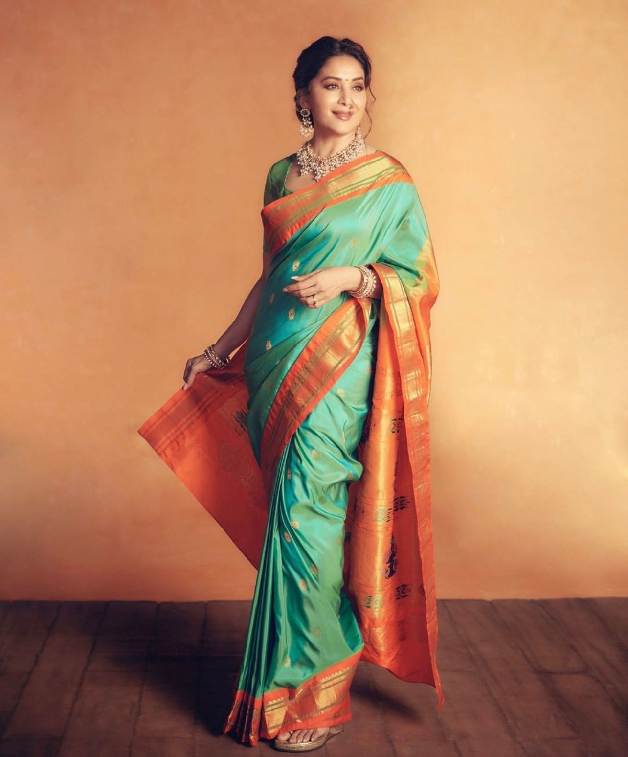 Luminous Soft Lichi Silk saree in teal/mint green with a wide, fiery orange contrast border featuring intricate temple zari weaving, gracefully worn by a smiling woman with traditional jewelry against an orange wall.