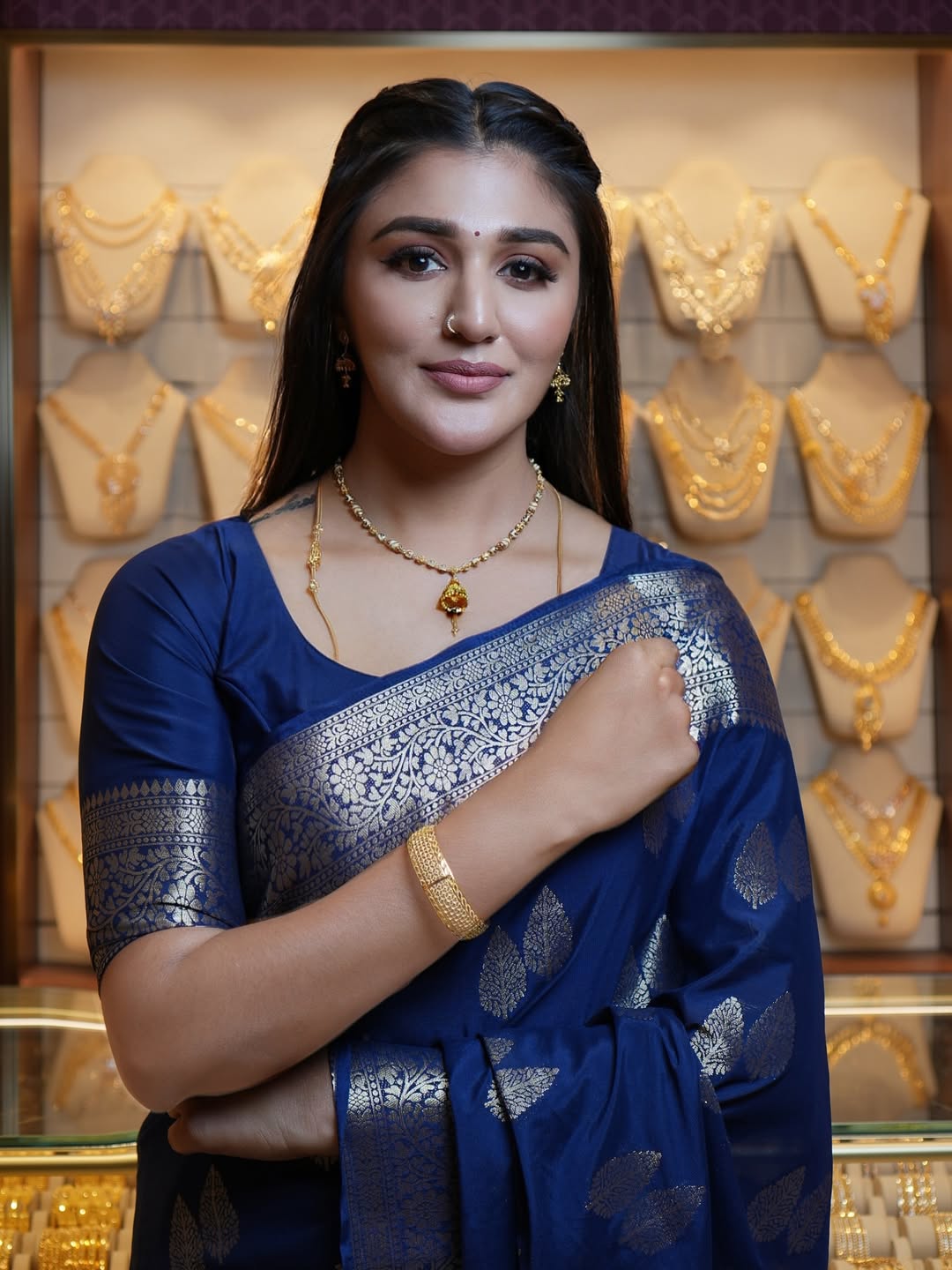 Woman wearing a blue saree with gold jewelry in front of a display of gold necklaces.