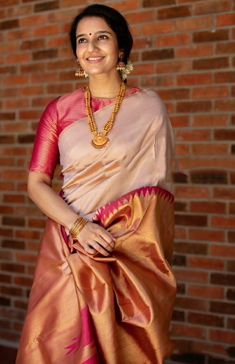 A smiling woman stands against a brick wall, elegantly draped in a soft Lichi silk saree in a blush golden-beige hue, featuring a rich rani pink and golden border, paired with a vibrant red blouse, gold temple jewelry, and gajra in her hair.