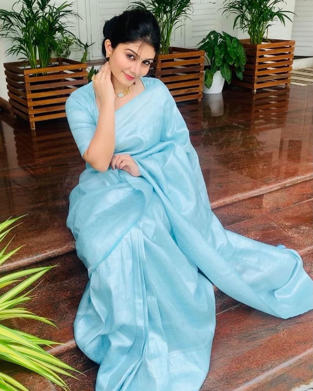A woman stands gracefully on a porch, holding a pink clutch and wearing a soft, shimmering aqua blue Lichi silk saree with a matching blouse, accessorized with a delicate gold necklace.