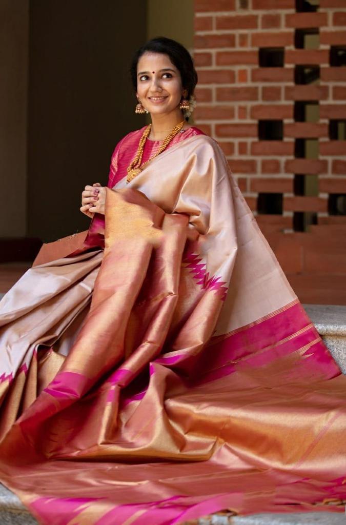 A smiling woman stands against a brick wall, elegantly draped in a soft Lichi silk saree in a blush golden-beige hue, featuring a rich rani pink and golden border, paired with a vibrant red blouse, gold temple jewelry, and gajra in her hair.