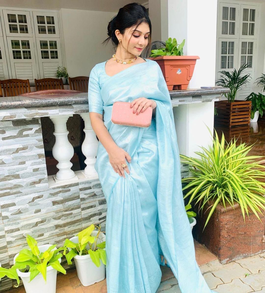 A woman stands gracefully on a porch, holding a pink clutch and wearing a soft, shimmering aqua blue Lichi silk saree with a matching blouse, accessorized with a delicate gold necklace.