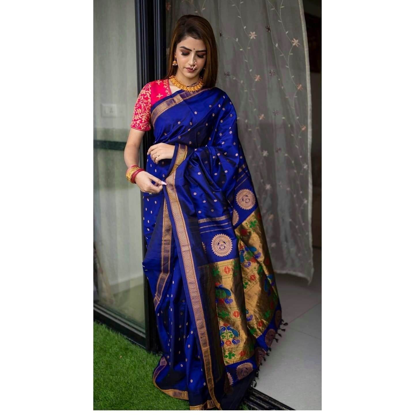 Exquisite Soft Lichi Silk saree in deep royal blue with gold circular motifs, featuring a magnificent multi-coloured Paithani-style pallu with intricate gold zari weaving, worn by a woman standing by a glass door