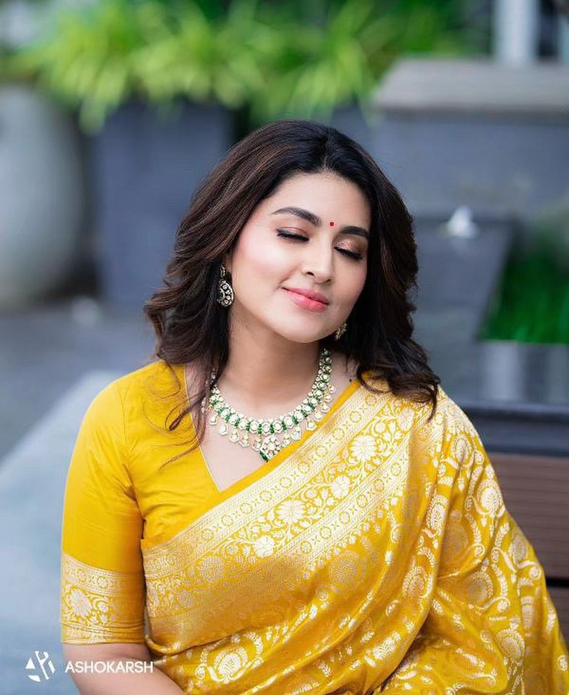 Woman in a yellow saree with green jewelry, sitting outdoors.