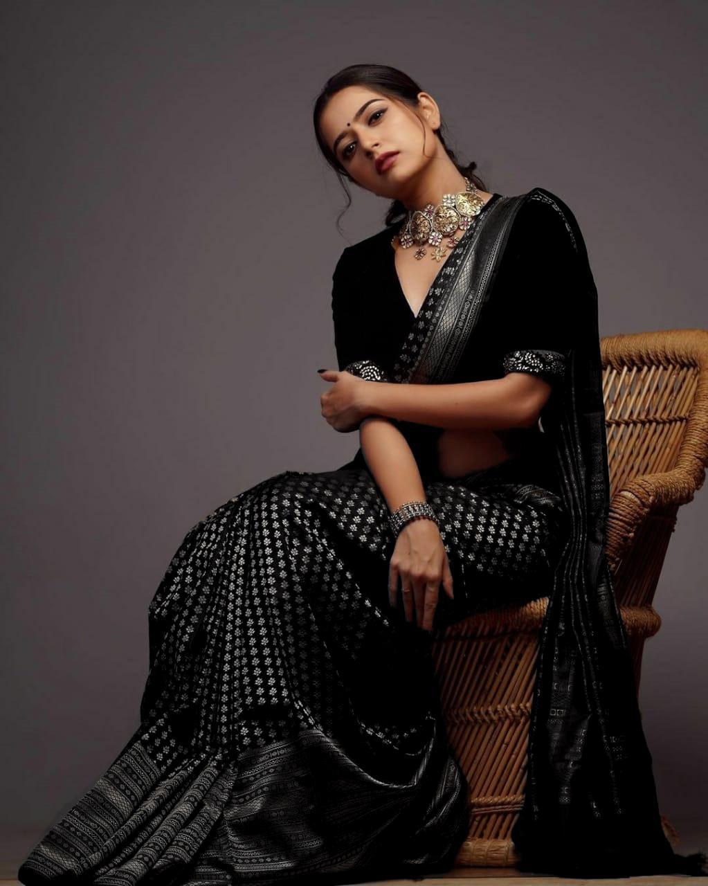 A woman models a black soft Lichi silk saree with silver patterned weaving, a matching velvet blouse, and traditional Indian jewelry, against a plain background.