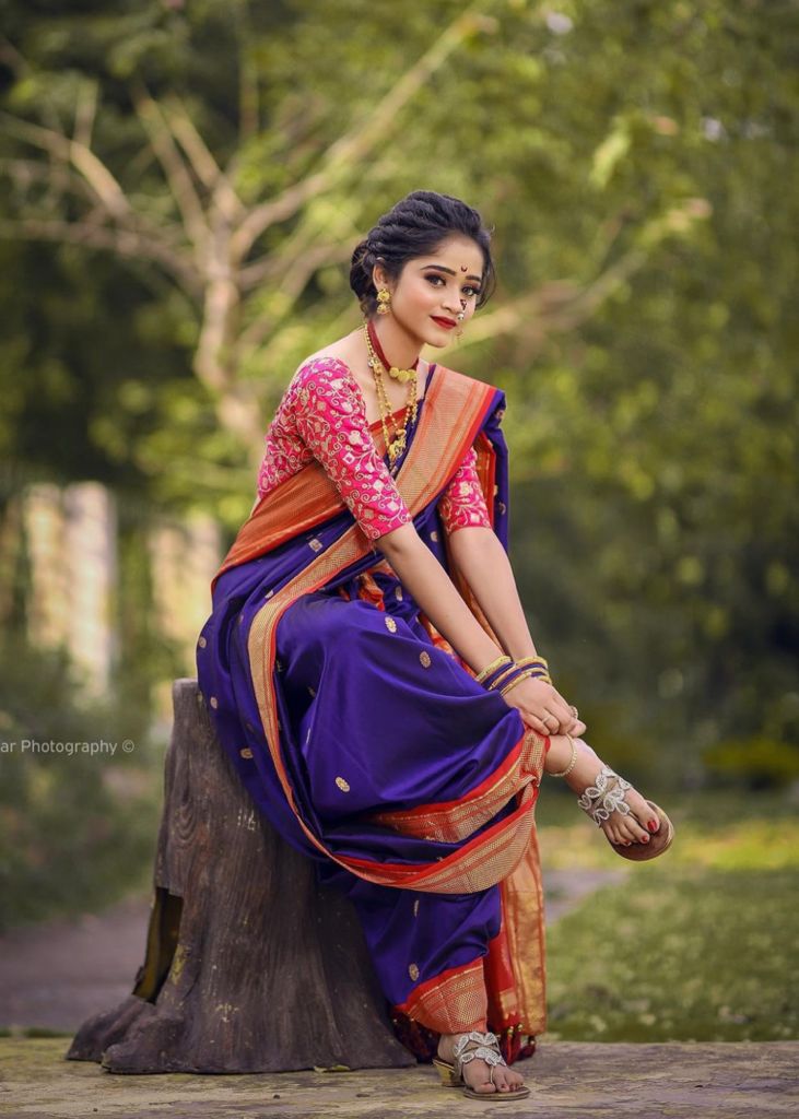 Deep amethyst purple Banarasi Soft Silk saree with scattered gold circular motifs and a bold red and gold zari woven border, beautifully styled on a woman with traditional Maharashtrian jewelry in an outdoor setting.