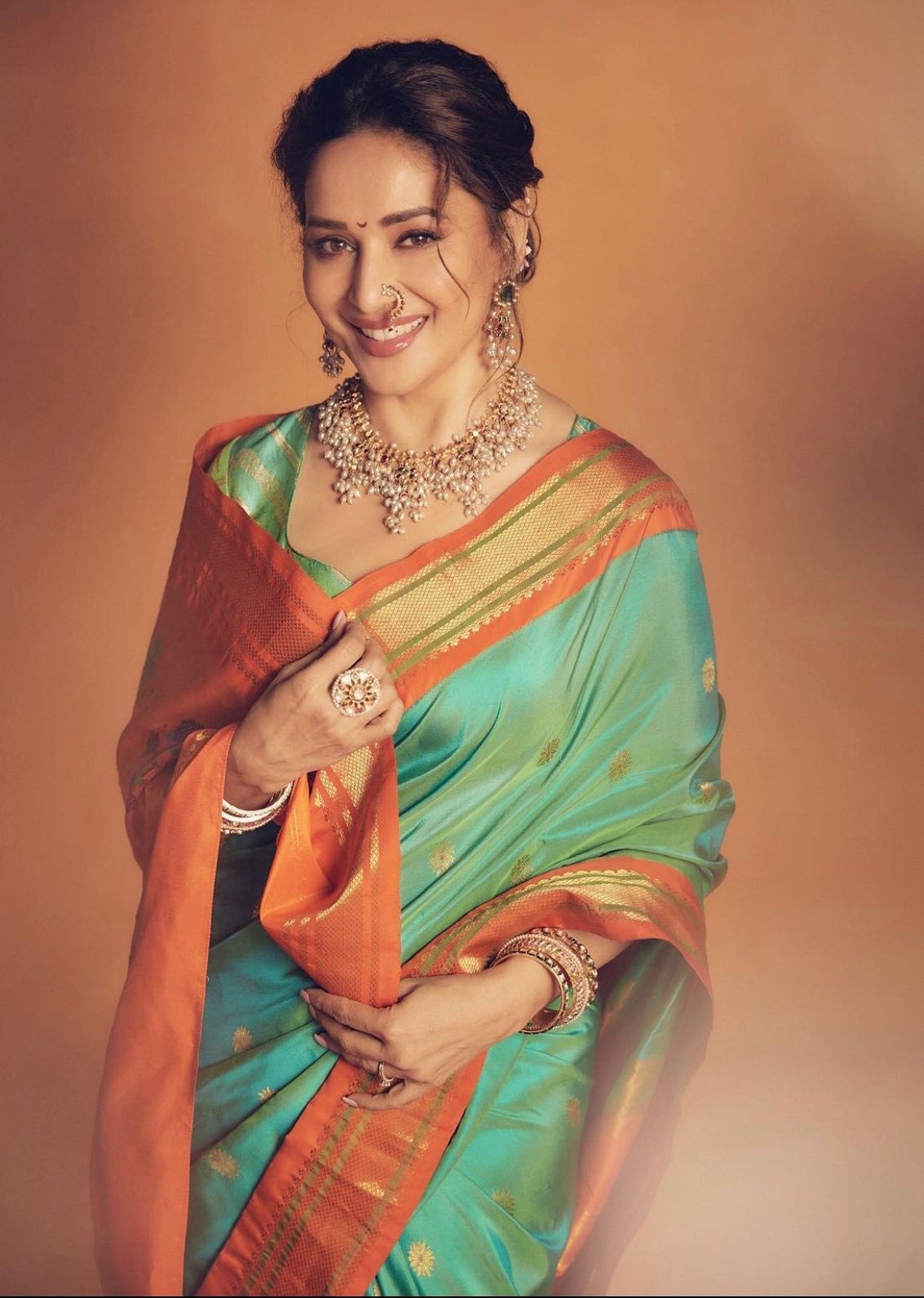 Luminous Soft Lichi Silk saree in teal/mint green with a wide, fiery orange contrast border featuring intricate temple zari weaving, gracefully worn by a smiling woman with traditional jewelry against an orange wall.