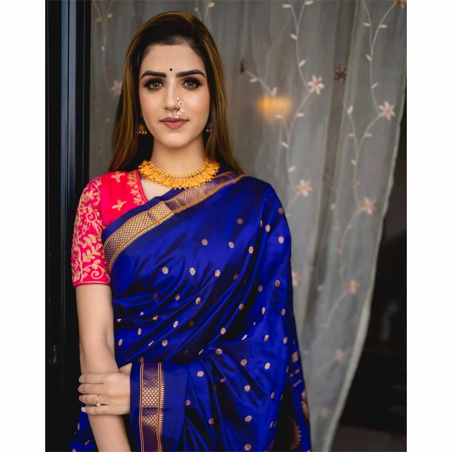 Exquisite Soft Lichi Silk saree in deep royal blue with gold circular motifs, featuring a magnificent multi-coloured Paithani-style pallu with intricate gold zari weaving, worn by a woman standing by a glass door