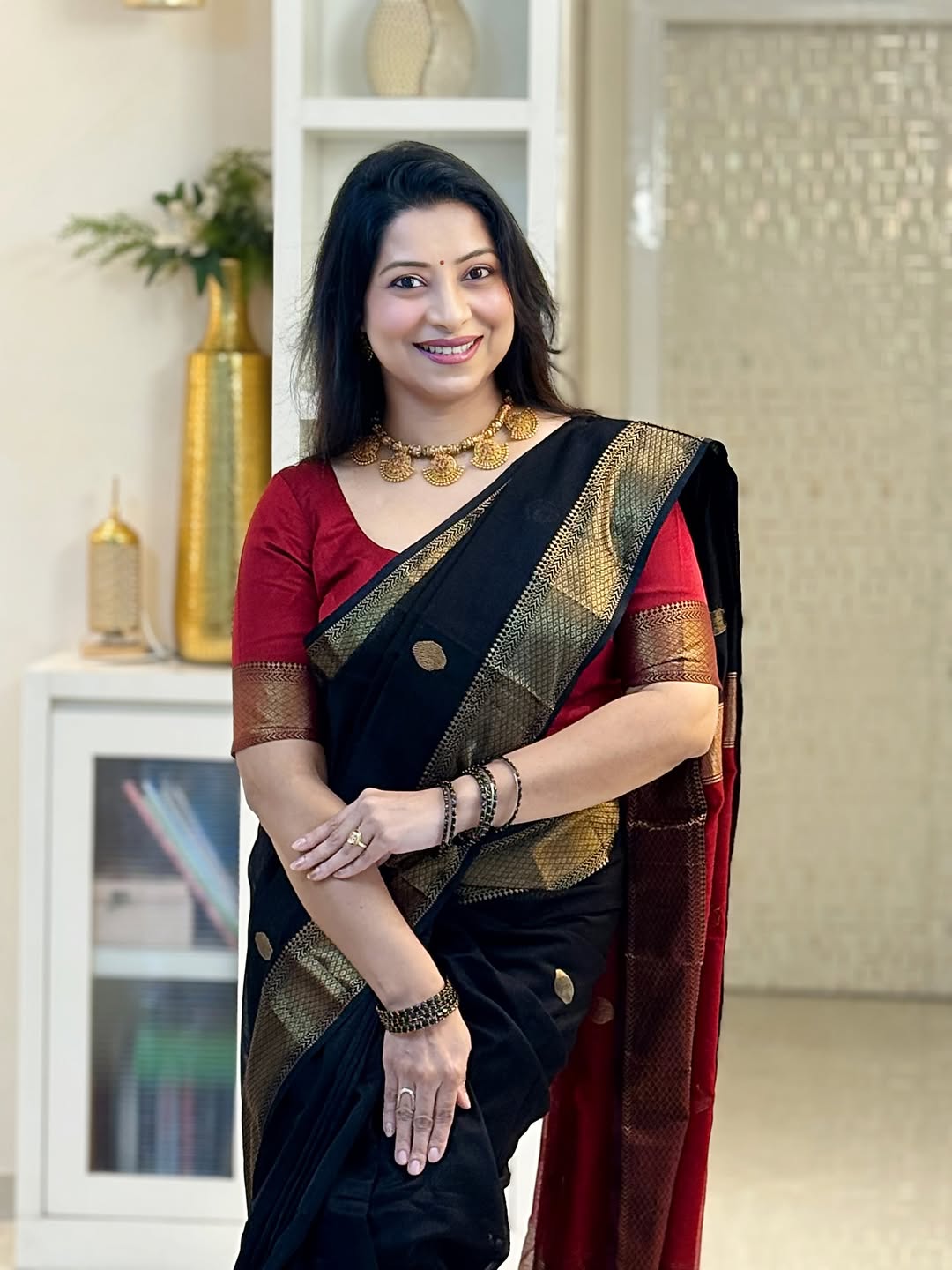 Woman in a black and red saree with gold accents standing indoors.