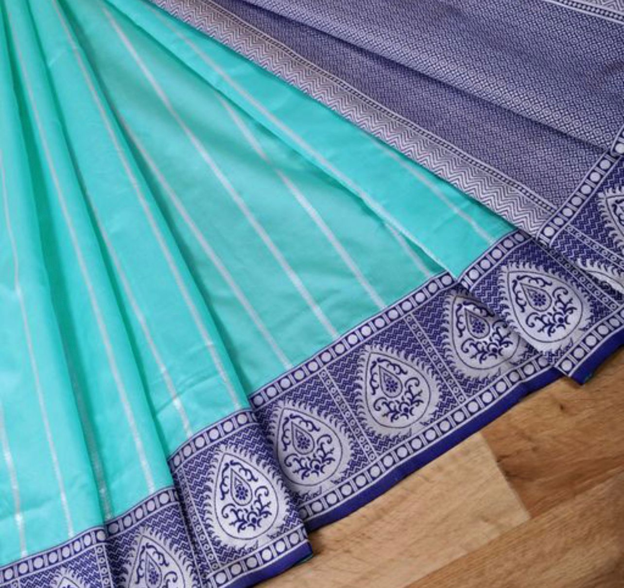 A smiling woman adorned in exquisite diamond and emerald jewelry, including a maang tikka and a layered necklace, wears a soft Lichi silk saree in a vibrant aqua blue with a contrasting silver and blue intricately woven border and pallu.