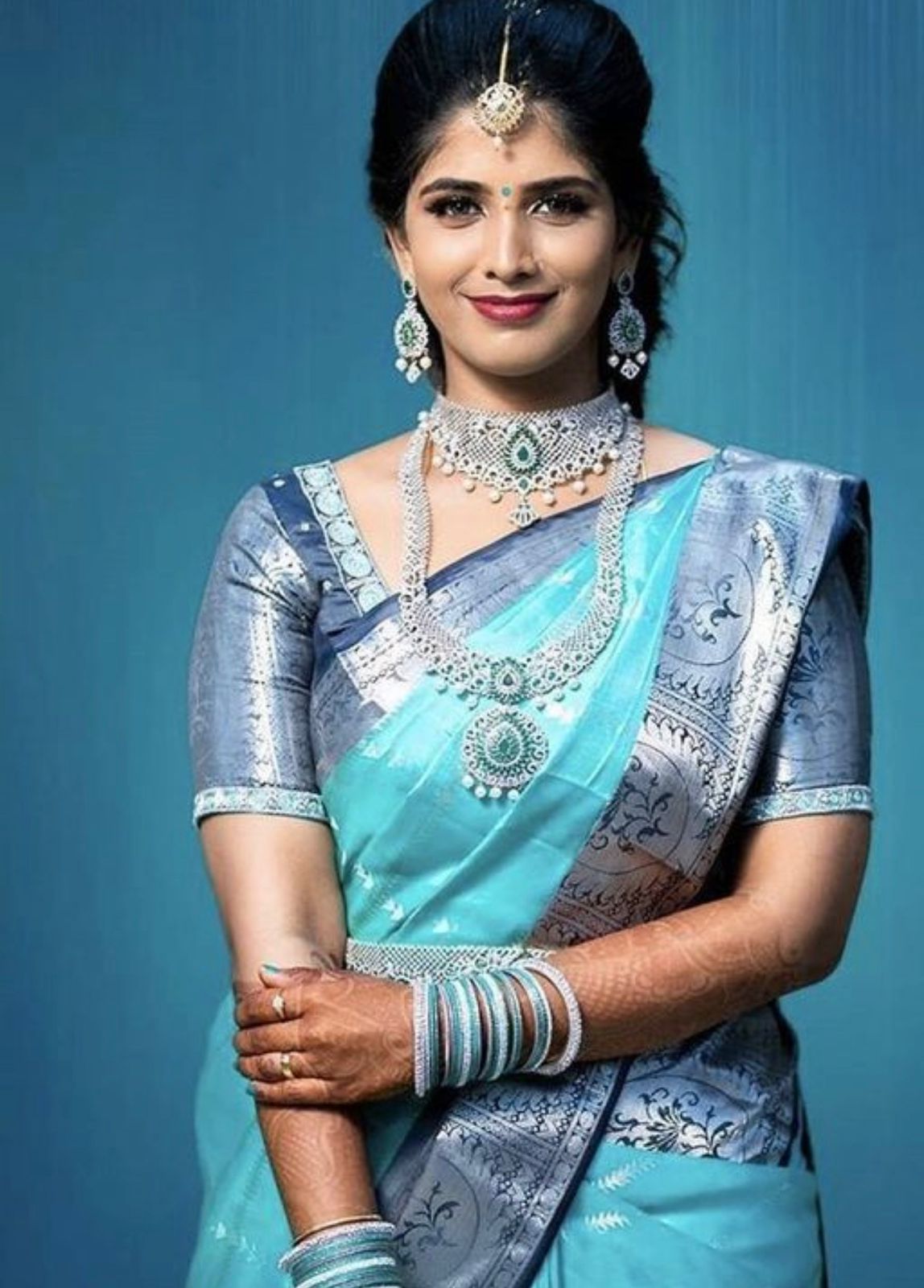 A smiling woman adorned in exquisite diamond and emerald jewelry, including a maang tikka and a layered necklace, wears a soft Lichi silk saree in a vibrant aqua blue with a contrasting silver and blue intricately woven border and pallu.