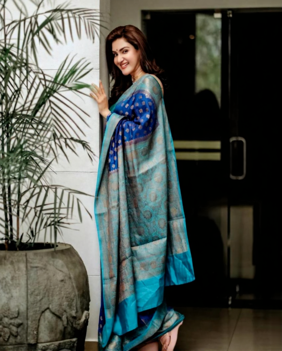 Royal blue Banarasi Silk saree with gold zari buttis and a teal contrast border, worn by a model in a natural light setting.