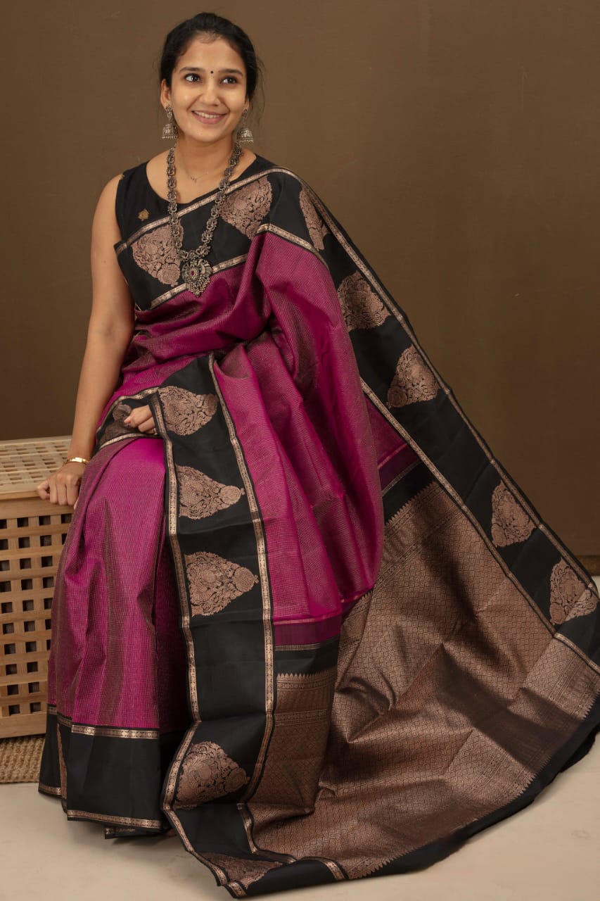Deep fuschia/magenta Banarasi Silk saree with black contrast panels on the pallu and border, adorned with bronze zari paisley motifs, draped on a woman sitting on a wooden stool indoors.