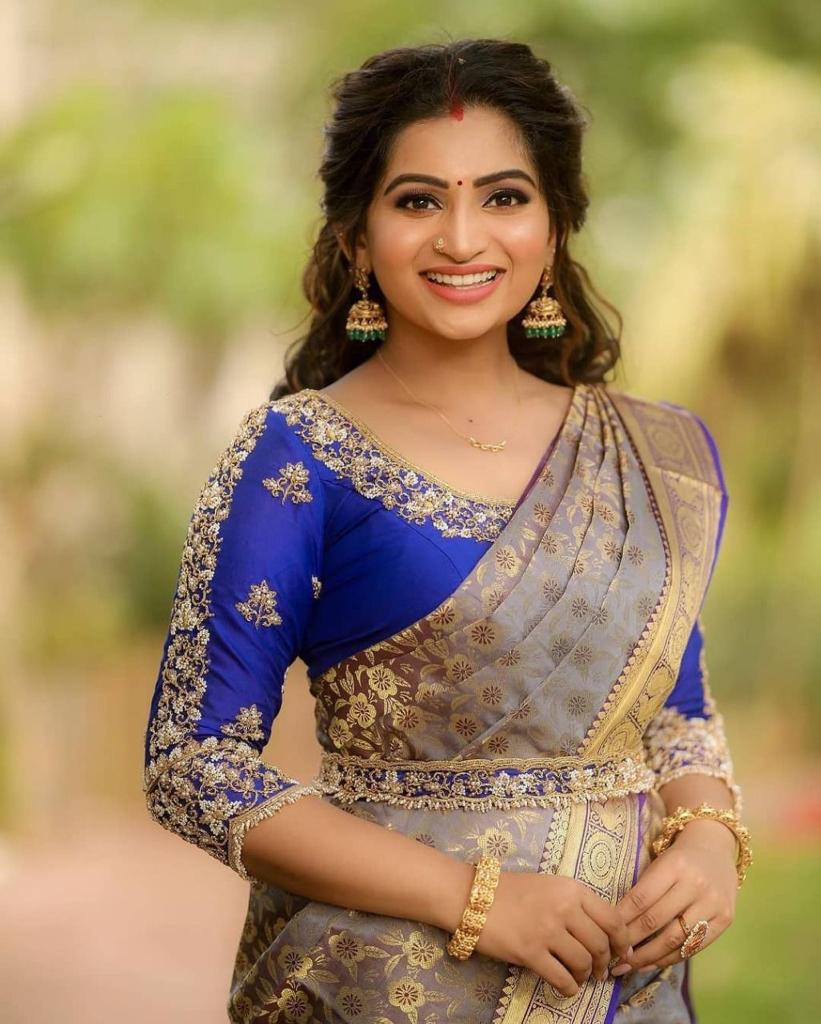 smiling woman sits outdoors, wearing a soft Lichi silk saree in elephant grey with gold floral patterns and a vibrant royal blue embroidered blouse, adorned with traditional jewelry.