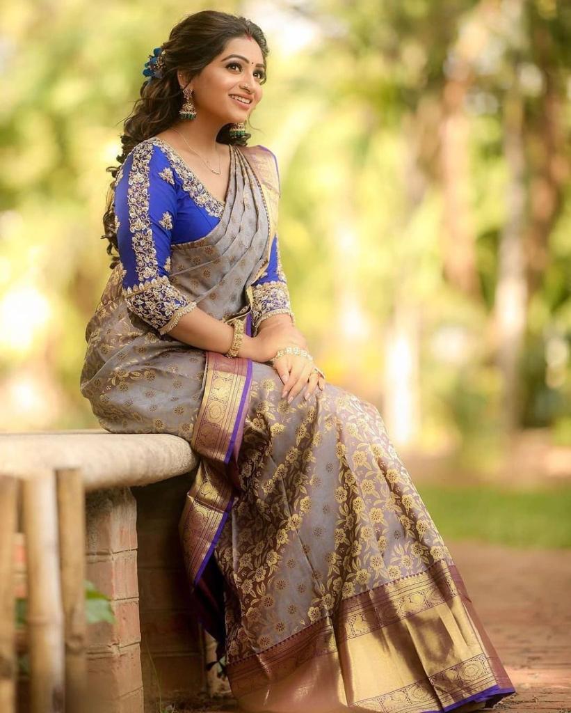 smiling woman sits outdoors, wearing a soft Lichi silk saree in elephant grey with gold floral patterns and a vibrant royal blue embroidered blouse, adorned with traditional jewelry.