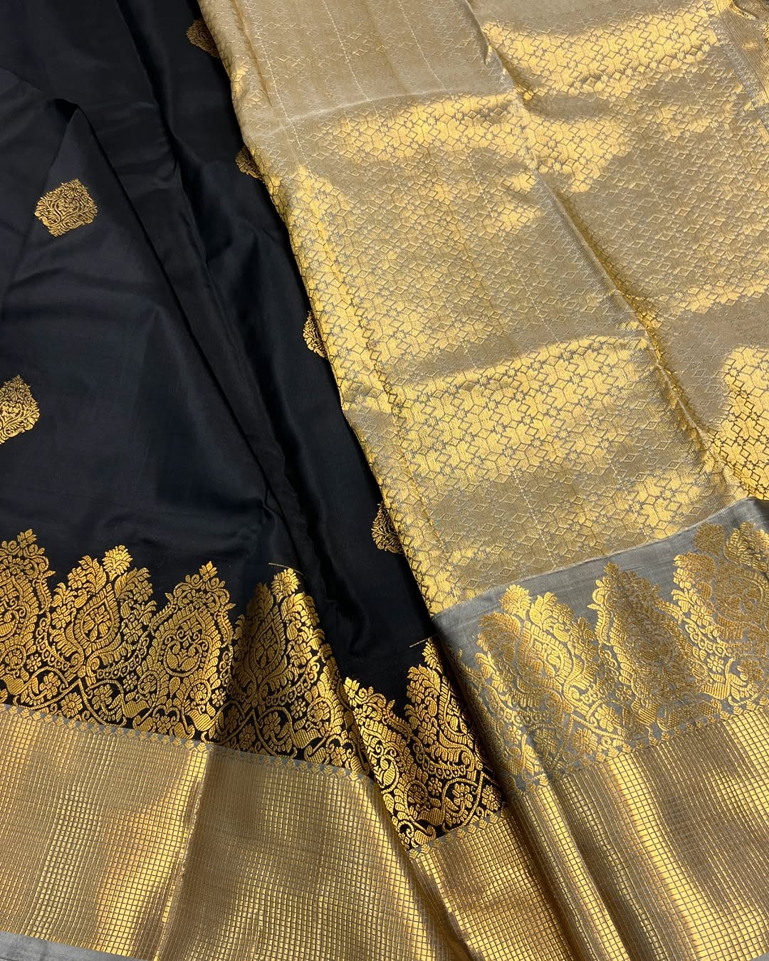 Classic jet black Banarasi Silk saree with scattered gold zari motifs, a wide intricately woven gold zari border, and a shimmering gold/silver zari contrast pallu, elegantly draped on a white textured surface.