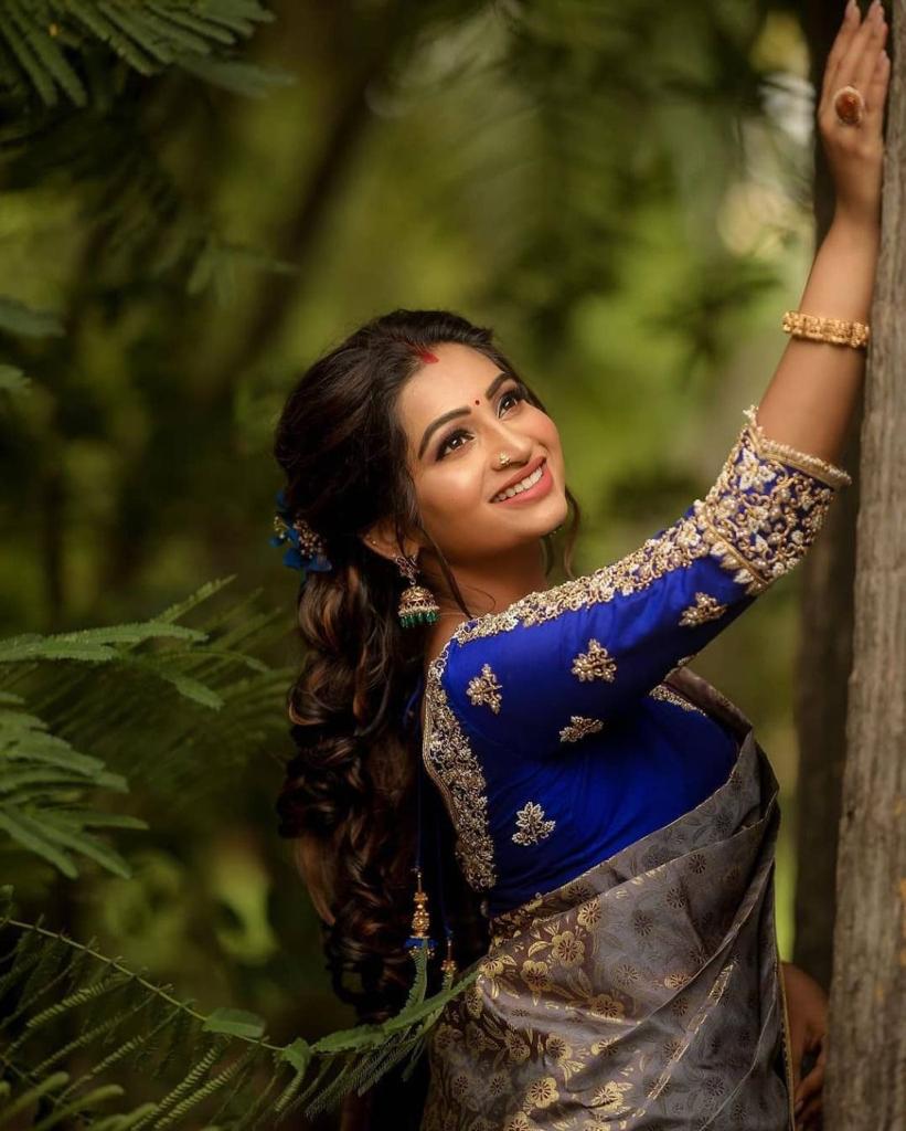 smiling woman sits outdoors, wearing a soft Lichi silk saree in elephant grey with gold floral patterns and a vibrant royal blue embroidered blouse, adorned with traditional jewelry.
