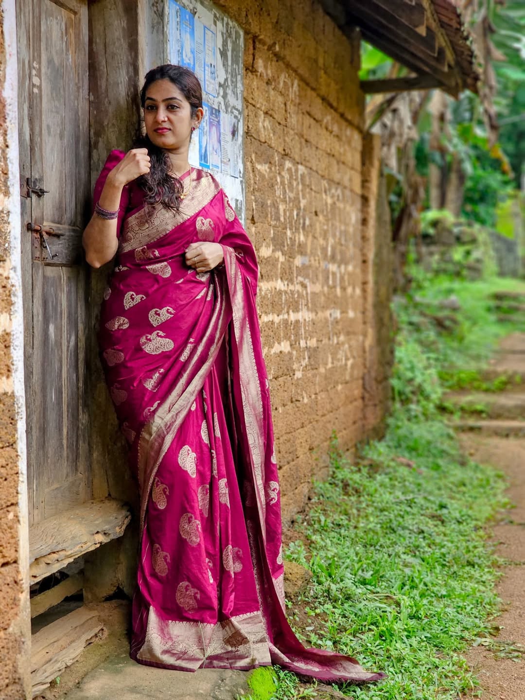 Woman in a pink saree standing in front of a rustic building with greenery around.