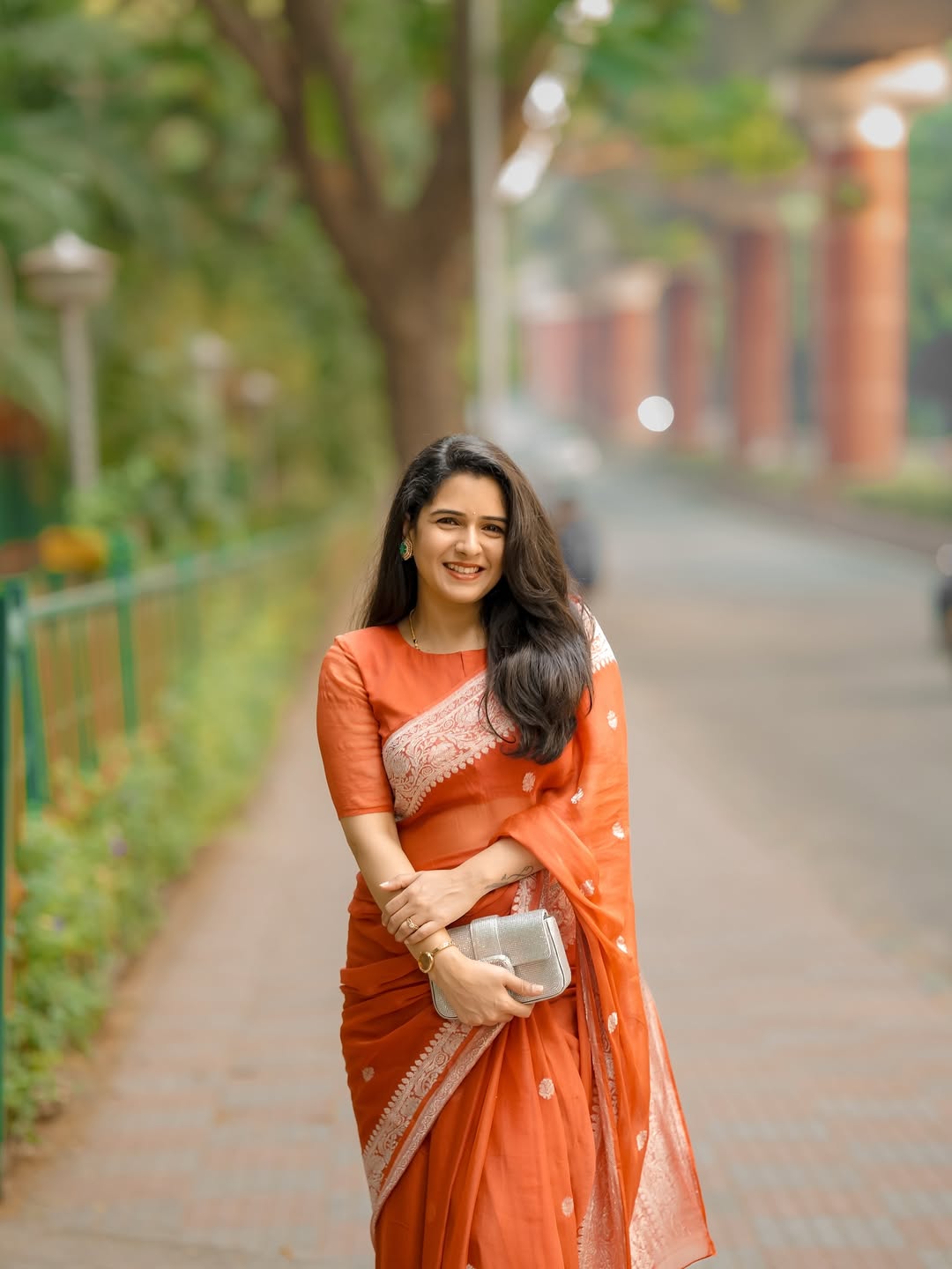 Woman in an orange saree walking on a path with greenery and a building in the background