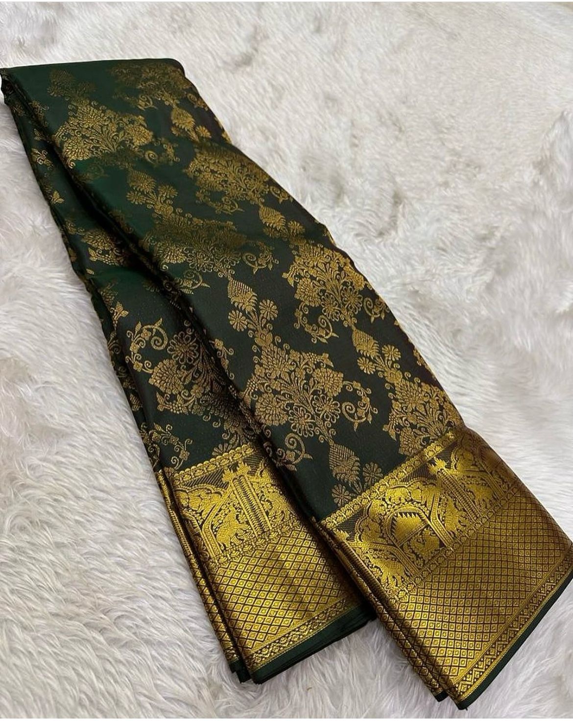 Deep forest green Banarasi Silk saree with dense all-over gold zari floral weaving and a wide border featuring traditional elephant motifs and geometric patterns, elegantly draped on a white textured surface.