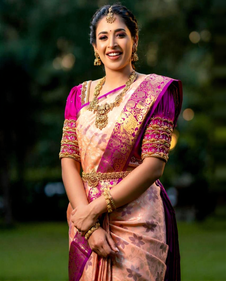 Radiant Banarasi Soft Silk saree in soft peach/cream with intricate floral zari motifs, featuring a vibrant fuchsia/magenta contrast border and heavy gold zari work, gracefully worn by a smiling woman with traditional jewelry.