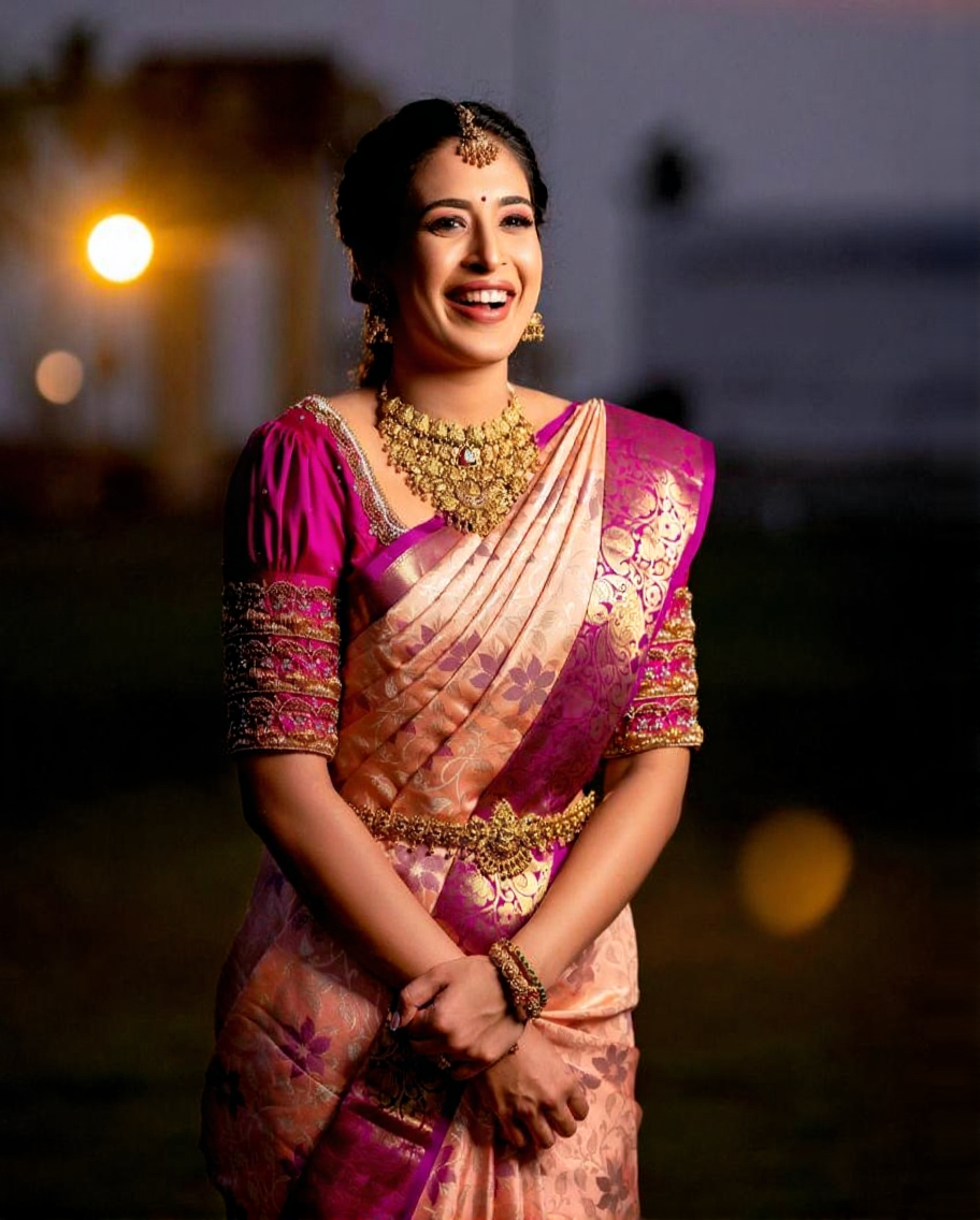 Radiant Banarasi Soft Silk saree in soft peach/cream with intricate floral zari motifs, featuring a vibrant fuchsia/magenta contrast border and heavy gold zari work, gracefully worn by a smiling woman with traditional jewelry.