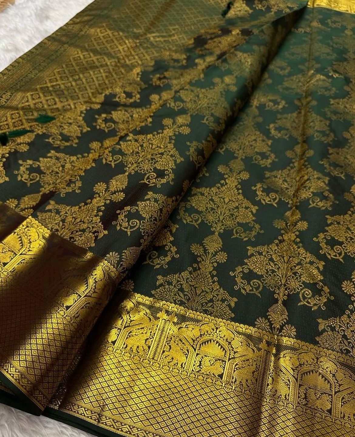 Deep forest green Banarasi Silk saree with dense all-over gold zari floral weaving and a wide border featuring traditional elephant motifs and geometric patterns, elegantly draped on a white textured surface.