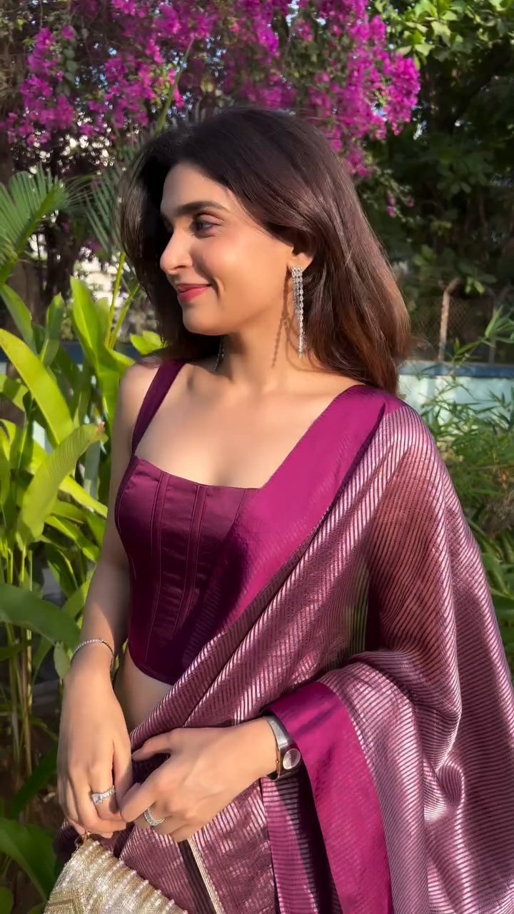 Woman in a purple saree standing among greenery with pink flowers.
