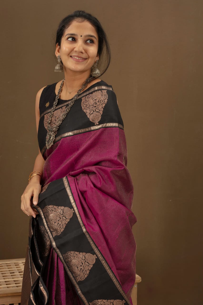 Deep fuschia/magenta Banarasi Silk saree with black contrast panels on the pallu and border, adorned with bronze zari paisley motifs, draped on a woman sitting on a wooden stool indoors.