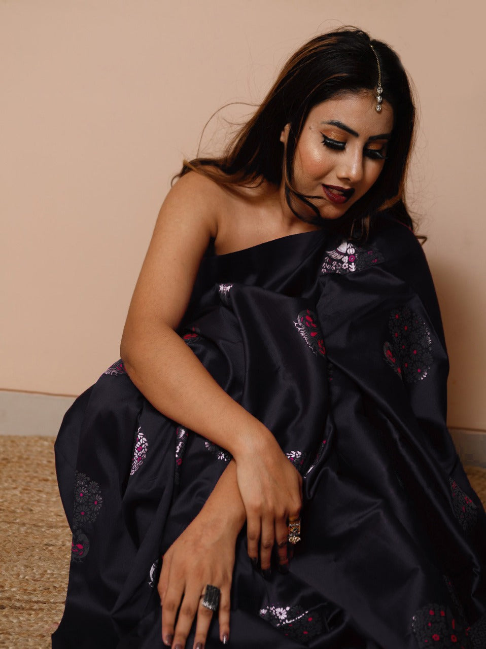 A woman sits on a woven mat against a light background, draped in a black soft Lichi silk saree featuring delicate silver and magenta floral patterns, with a single maang tikka adorning her forehead.