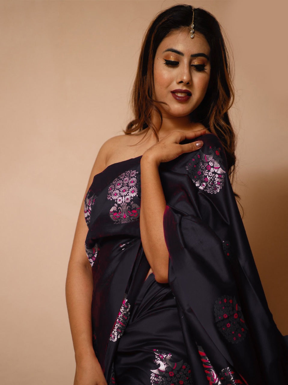 A woman sits on a woven mat against a light background, draped in a black soft Lichi silk saree featuring delicate silver and magenta floral patterns, with a single maang tikka adorning her forehead.