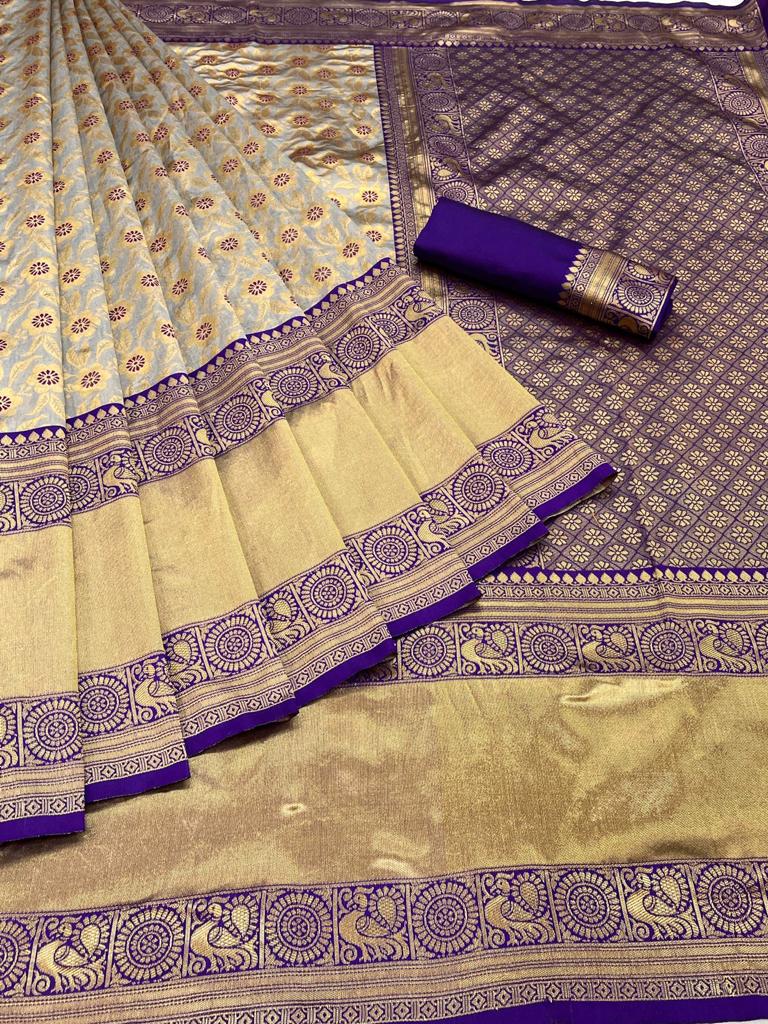 smiling woman sits outdoors, wearing a soft Lichi silk saree in elephant grey with gold floral patterns and a vibrant royal blue embroidered blouse, adorned with traditional jewelry.