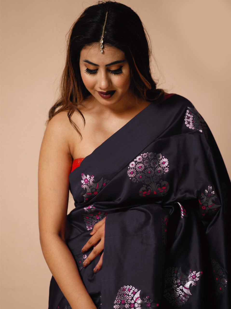 A woman sits on a woven mat against a light background, draped in a black soft Lichi silk saree featuring delicate silver and magenta floral patterns, with a single maang tikka adorning her forehead.