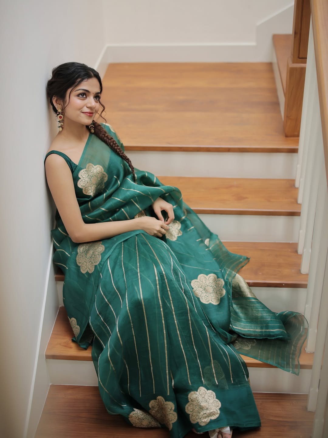 Woman in a green saree with floral patterns sitting on wooden stairs.
