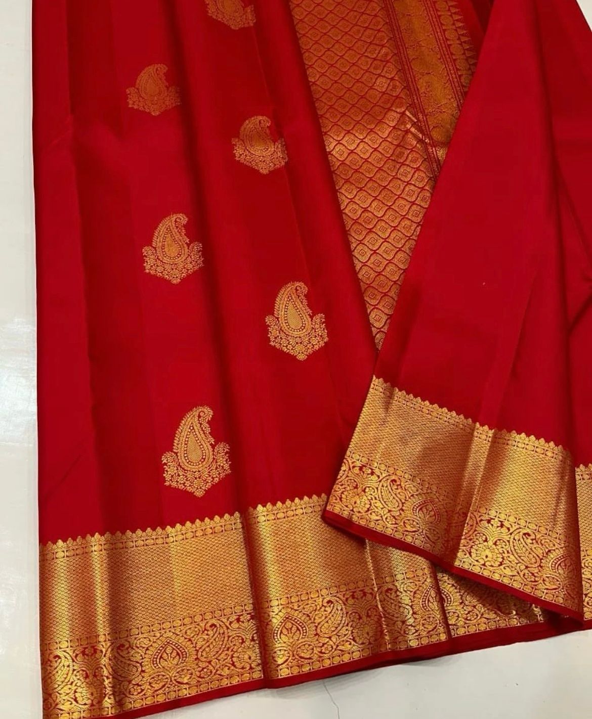 Vibrant rich red Banarasi Silk saree with scattered gold zari mango (paisley) buttas and a wide, intricately woven gold zari border and pallu, elegantly draped on a white surface.