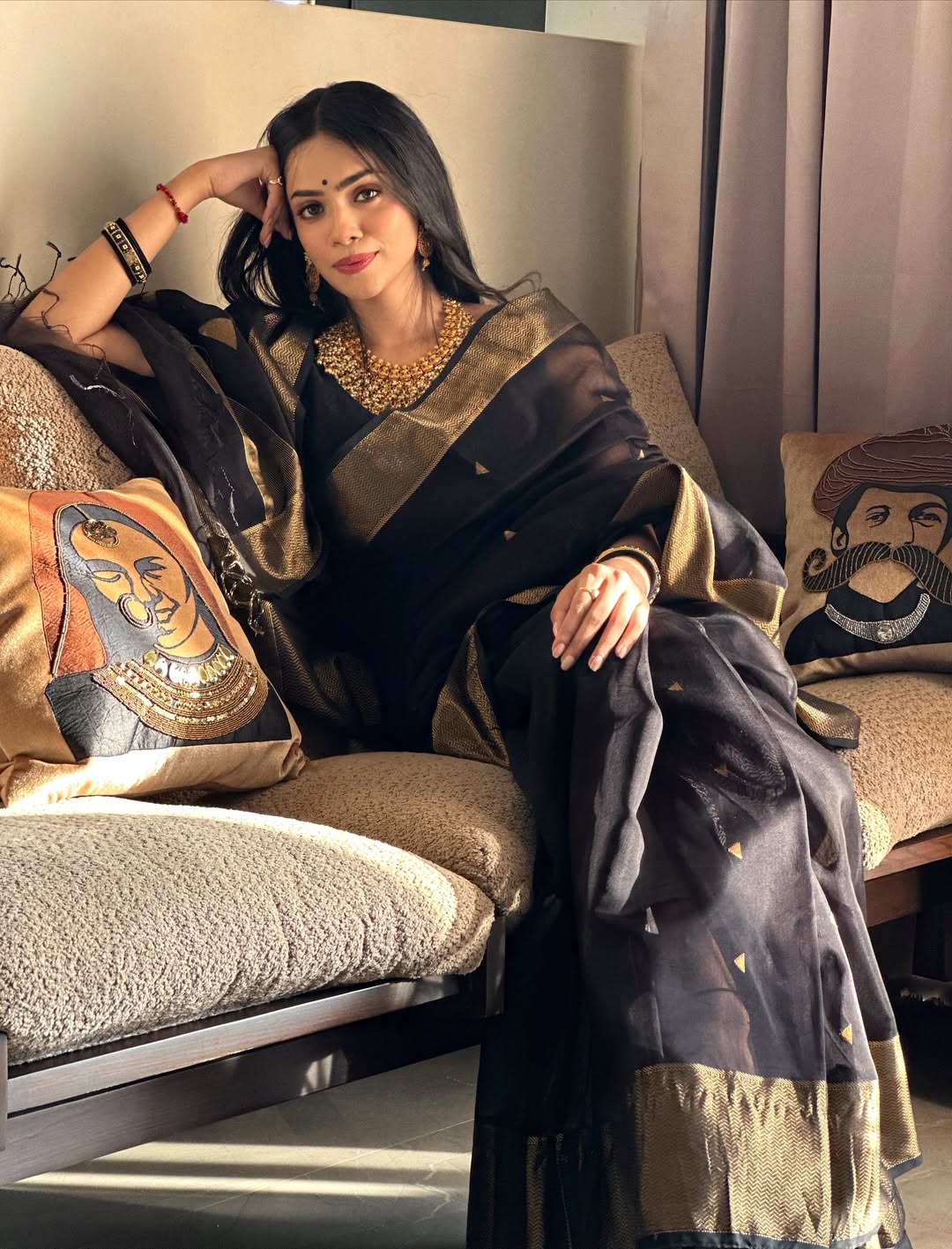 Woman in a black and gold saree sitting on a couch with decorative pillows.