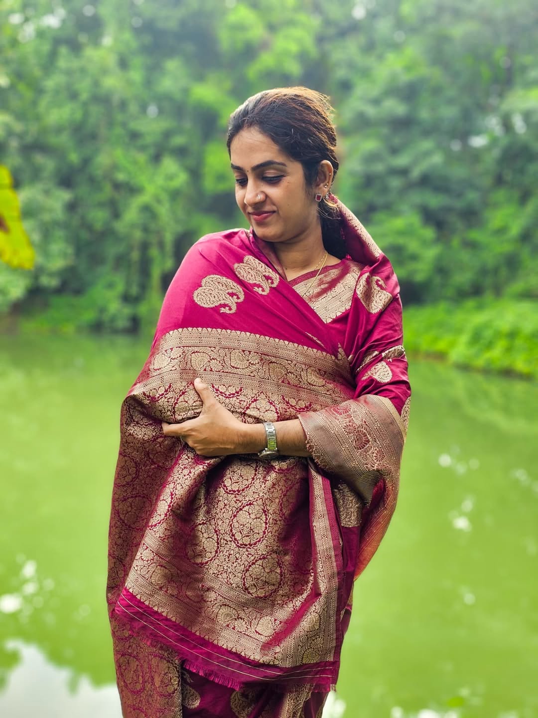 Woman in a pink and gold saree standing in front of a green, blurred natural background