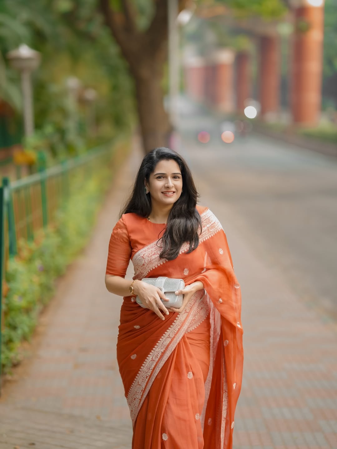 Woman in an orange saree standing on a pathway with greenery and a building in the background