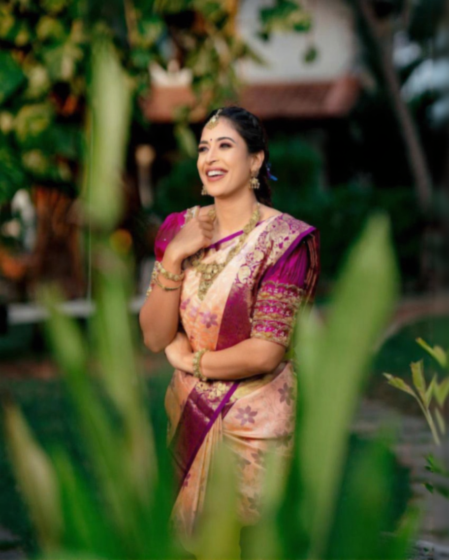Radiant Banarasi Soft Silk saree in soft peach/cream with intricate floral zari motifs, featuring a vibrant fuchsia/magenta contrast border and heavy gold zari work, gracefully worn by a smiling woman with traditional jewelry.